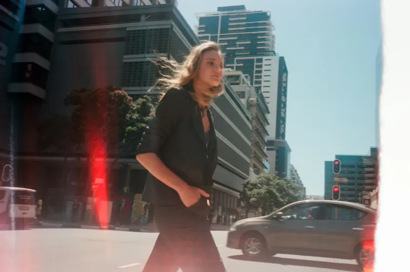 Blonde woman in black blazer and pants strides confidently on urban street amid skyscrapers and traffic.