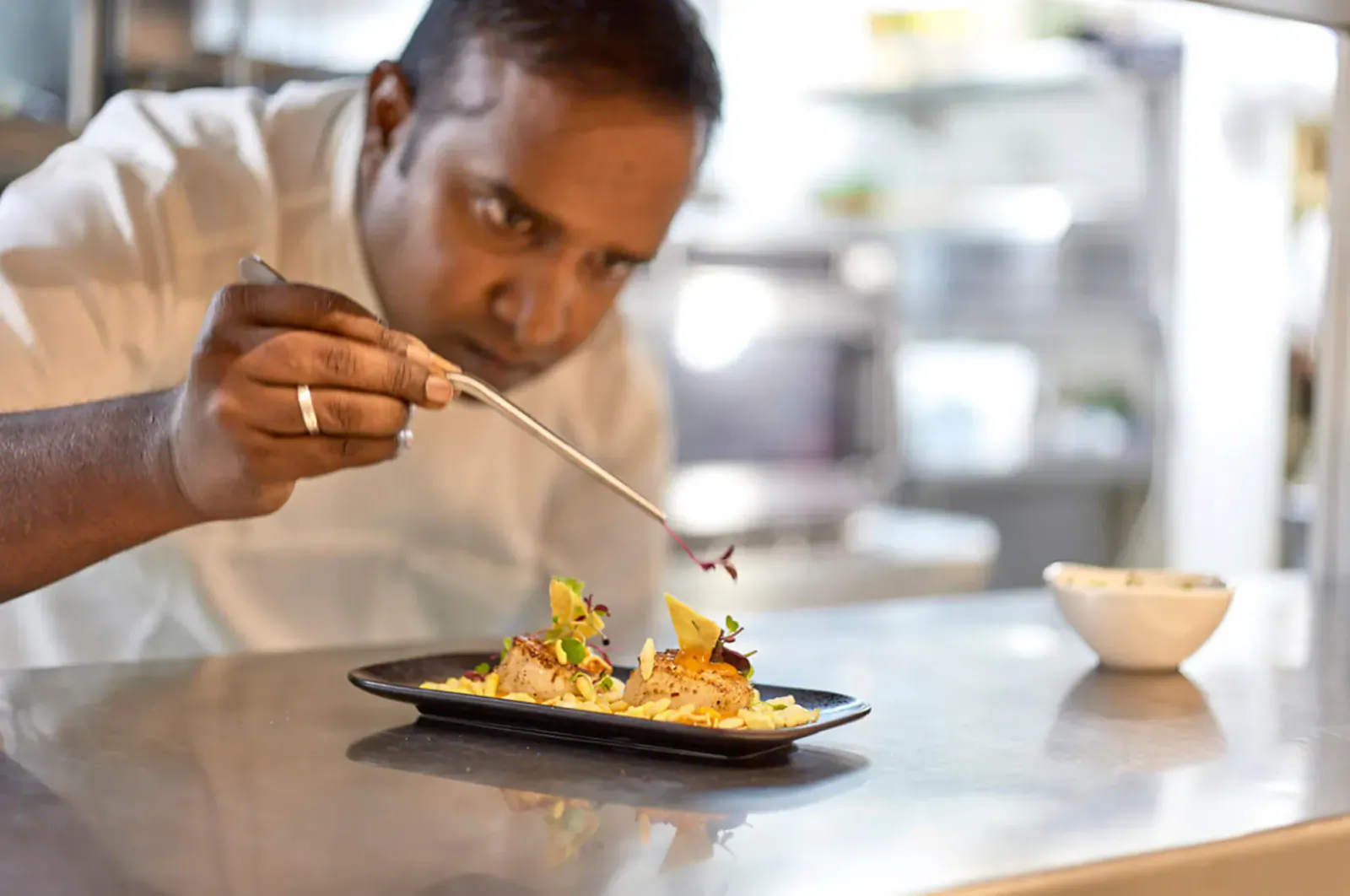 Chef in white uniform garnishing scallop dish with tweezers on black plate in restaurant kitchen