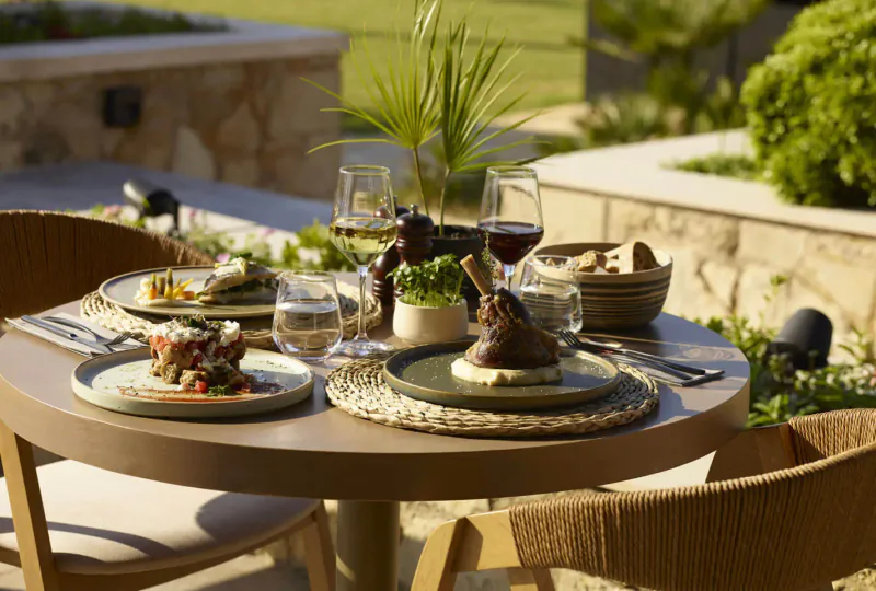 Outdoor table at Asterion Suites & Spa, Crete: plates of roasted lamb, cheese, veggies, bread, wine glasses amid greenery.