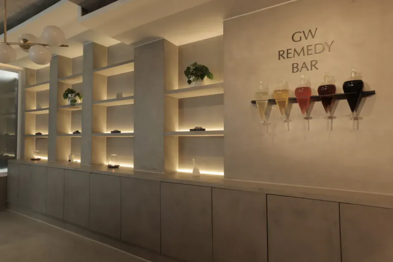 GW Remedy Bar in Grey Wolfe London wellness space: modern white shelves with plants, backlit cabinets, and six colorful remedy-filled glasses on wall.