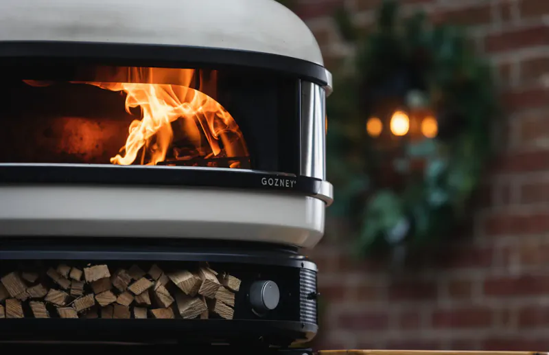 Gozney wood-fired pizza oven with blazing flames inside, wood stack below, Christmas wreath and brick wall backdrop