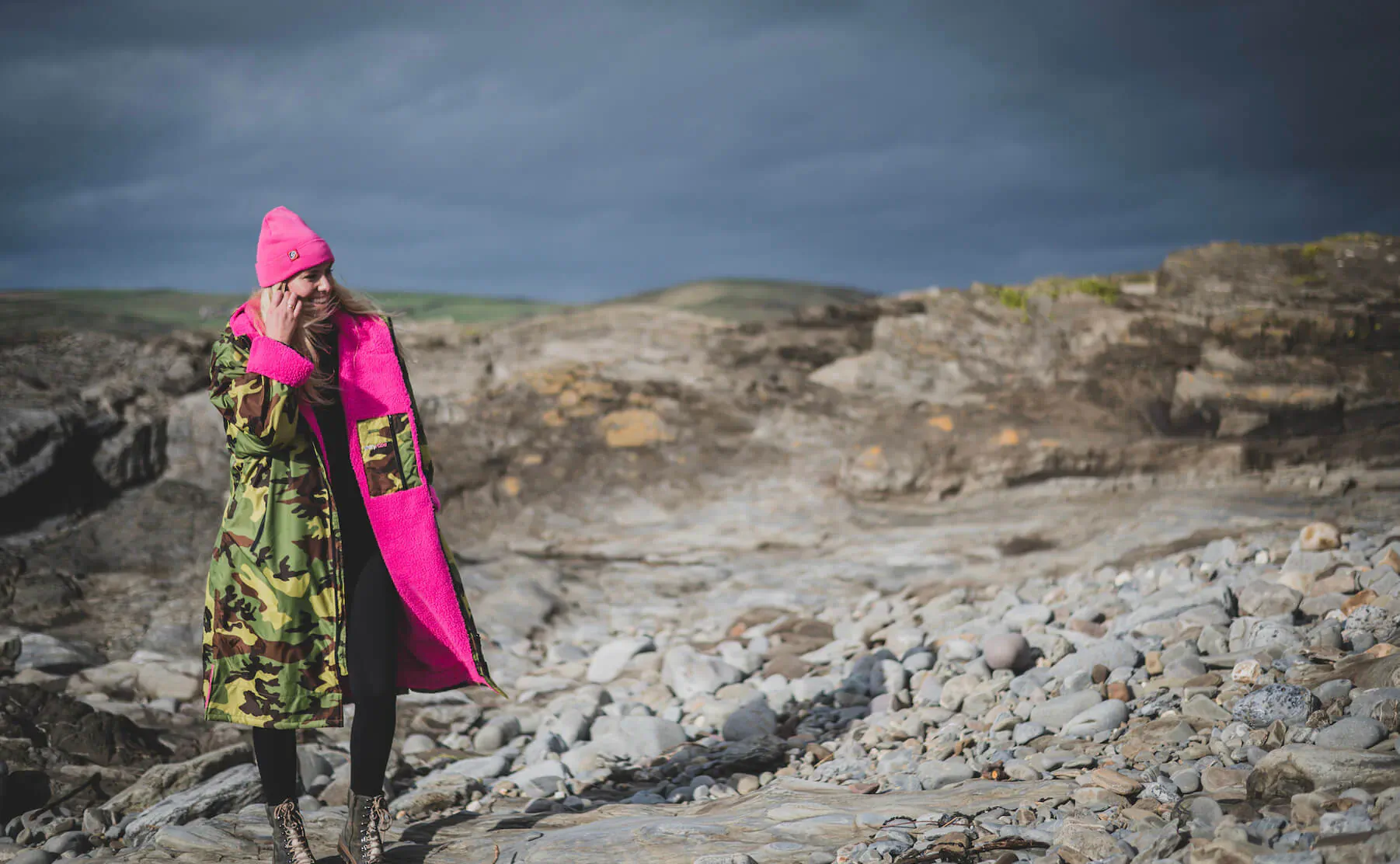 Woman in pink beanie, camouflage dryrobe with pink lining, and black leggings stands on rocky coastal cliffs under cloudy sky