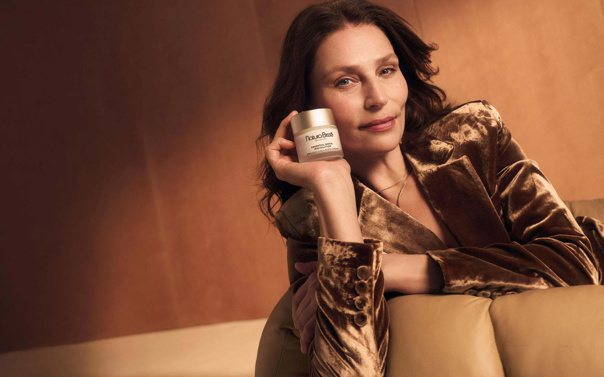 Woman in bronze velvet blazer holds white skincare jar, smiling on tan sofa against warm wall