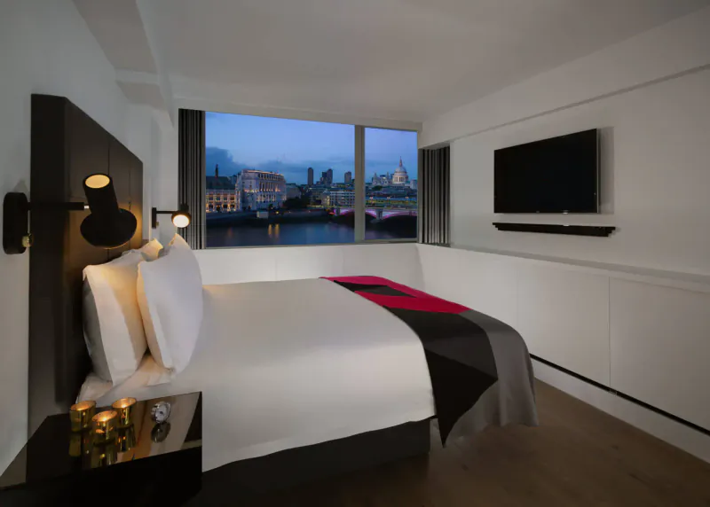 Modern hotel room with king bed, wall lamp, TV, and panoramic twilight view of River Thames and city skyline.