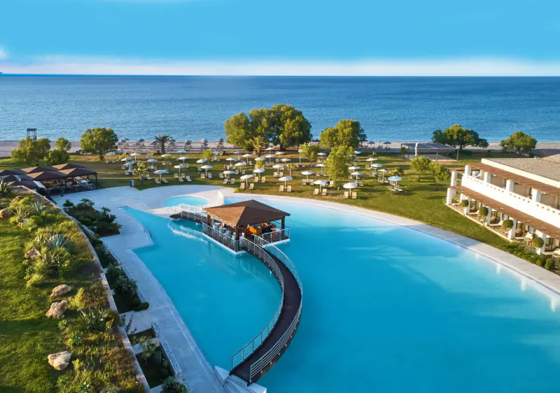 Aerial view of Cavo Spada resort's turquoise lagoon pool with central tiki bar, lounge chairs, greenery, and sea beyond in Crete.