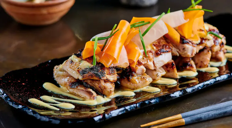 Grilled chicken skewers topped with carrot batons and green onions on black plate with sauce and chopsticks