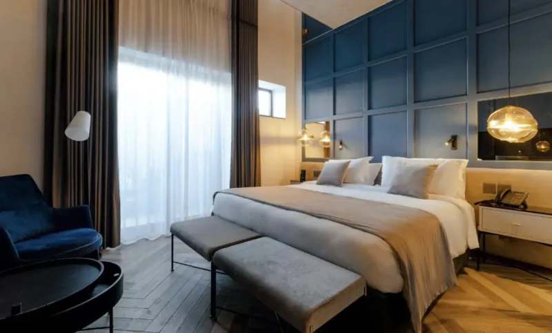 Elegant Embassy Valletta Hotel bedroom with navy accent wall, king bed, velvet chair, lamps, and large window.
