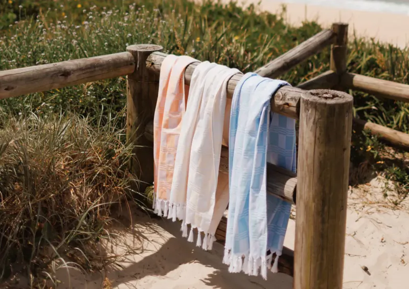 Assorted fringed beach towels in orange, white, and blue draped over a wooden fence by sandy grass.