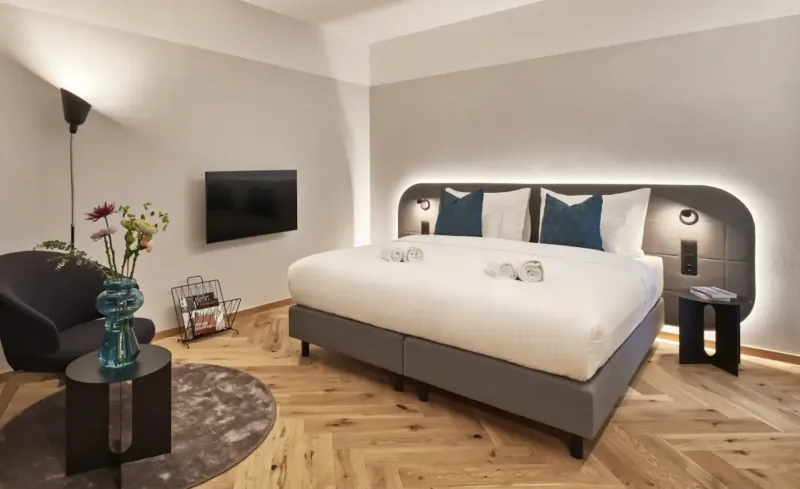 Modern Vienna hotel room with king bed, teal pillows, wall-mounted TV, vase of flowers, and bedside lamps.