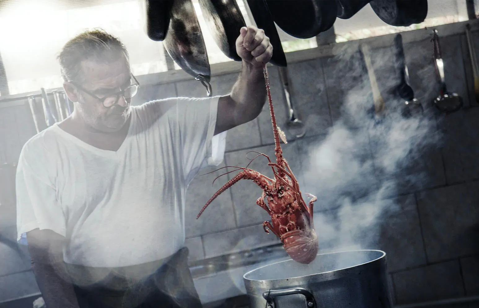 Chef in white shirt holding red lobster over steaming pot in kitchen with hanging utensils