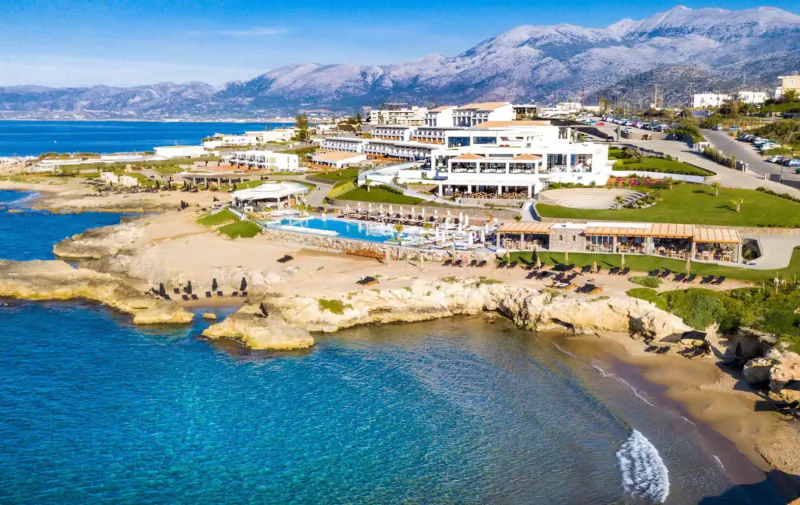 Aerial view of Abaton Island Resort & Spa, white buildings, infinity pool, beach, and sea with mountains backdrop.