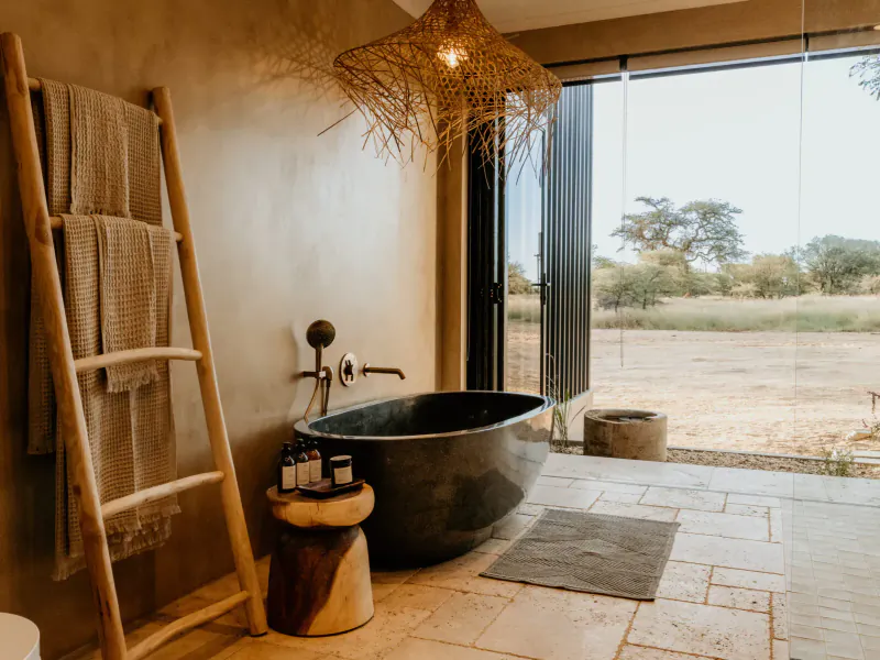 Luxurious savanna villa bathroom with black freestanding tub, rattan lamp, towels on ladder, and expansive wilderness view.