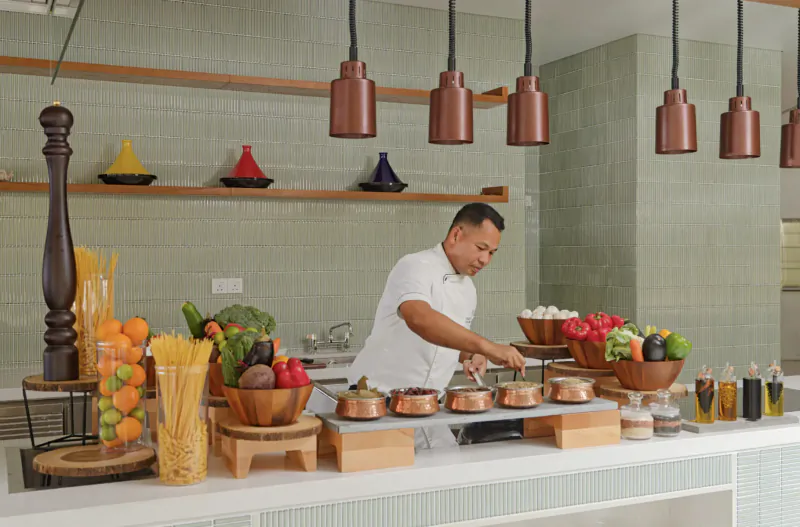 Chef in white uniform preparing vegetables on buffet counter with copper pots, fruits, and Moroccan lanterns at Amari Raaya Maldives