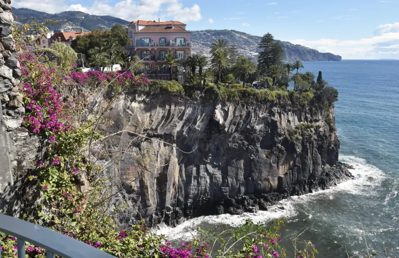 Les Suites Hotel at The Cliff Bay perched on lush cliffs overlooking the ocean in Funchal, Madeira, with purple bougainvillea and palm trees.