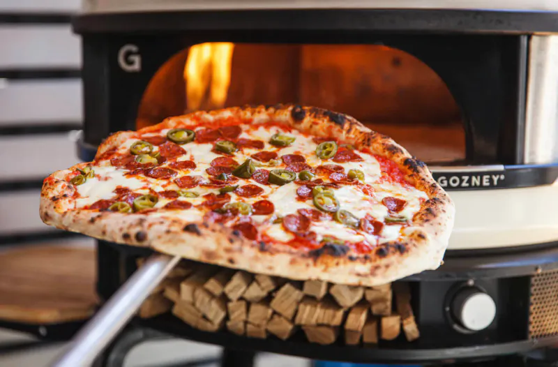 Pizza peel holding pepperoni pizza from lit Gozney oven, wood stacked nearby.