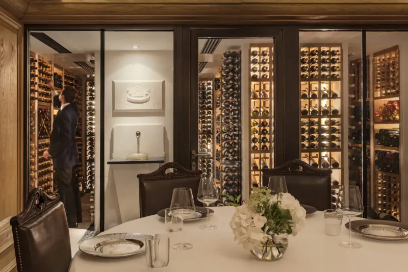 Elegant wine cellar with wooden racks, man in suit browsing bottles, dining table set with wine glasses and white flowers