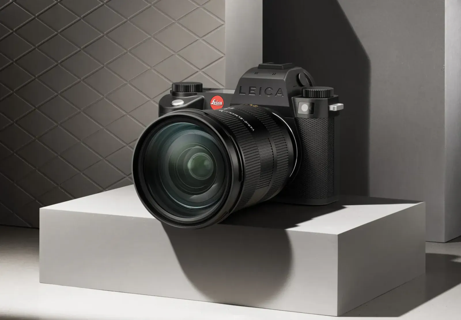 Black Leica SL3-S camera with large lens on white pedestal against gray quilted backdrop