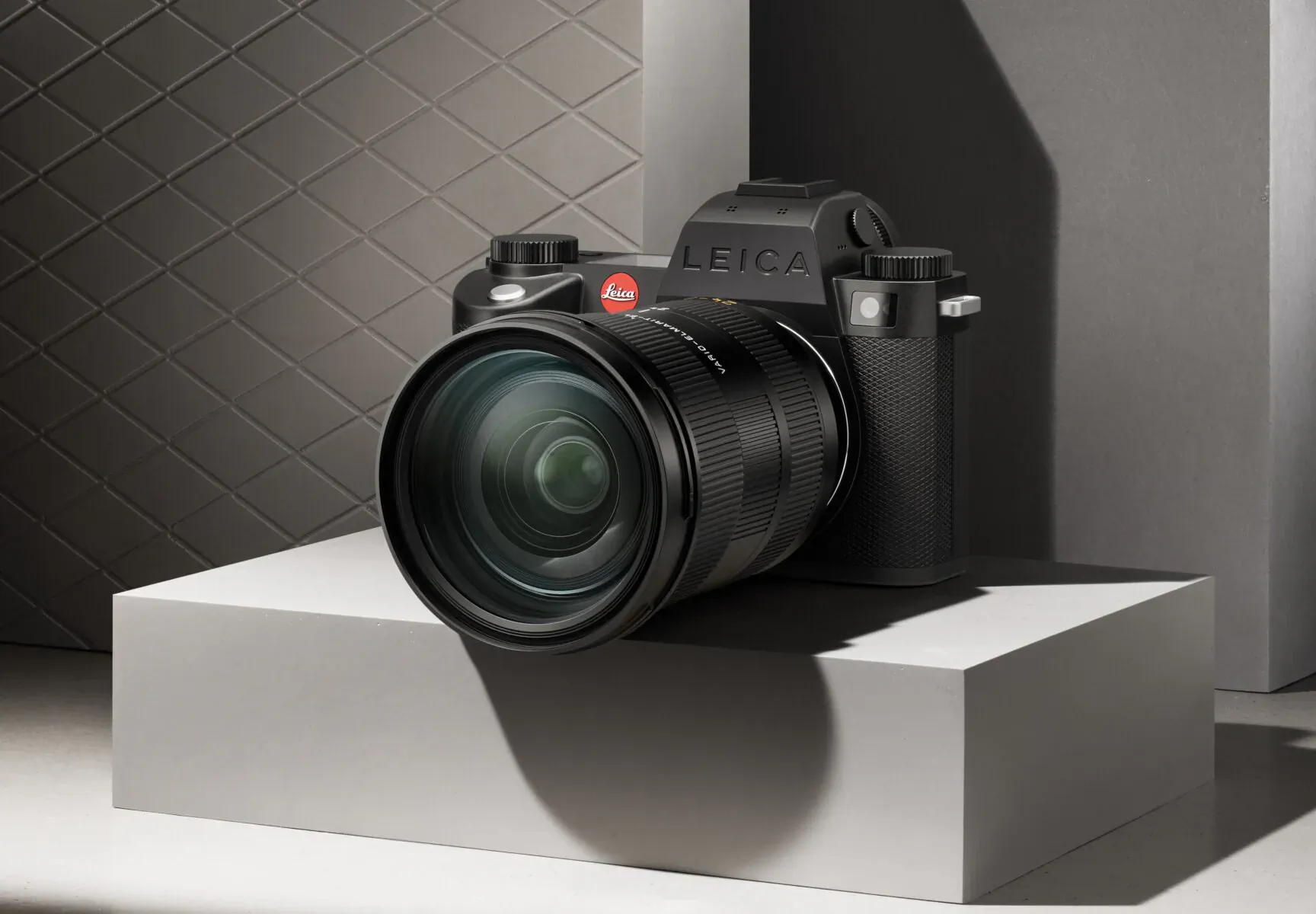 Black Leica SL3-S camera with large lens on white pedestal against gray quilted backdrop