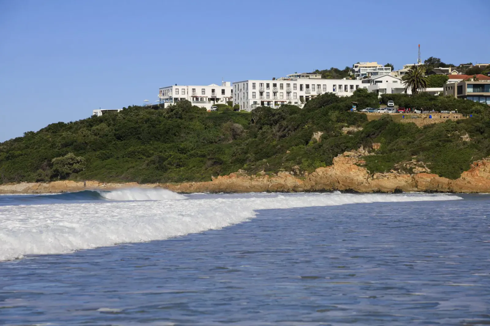 The Plettenberg Boutique Hotel on green hillside overlooking ocean waves, clear blue sky