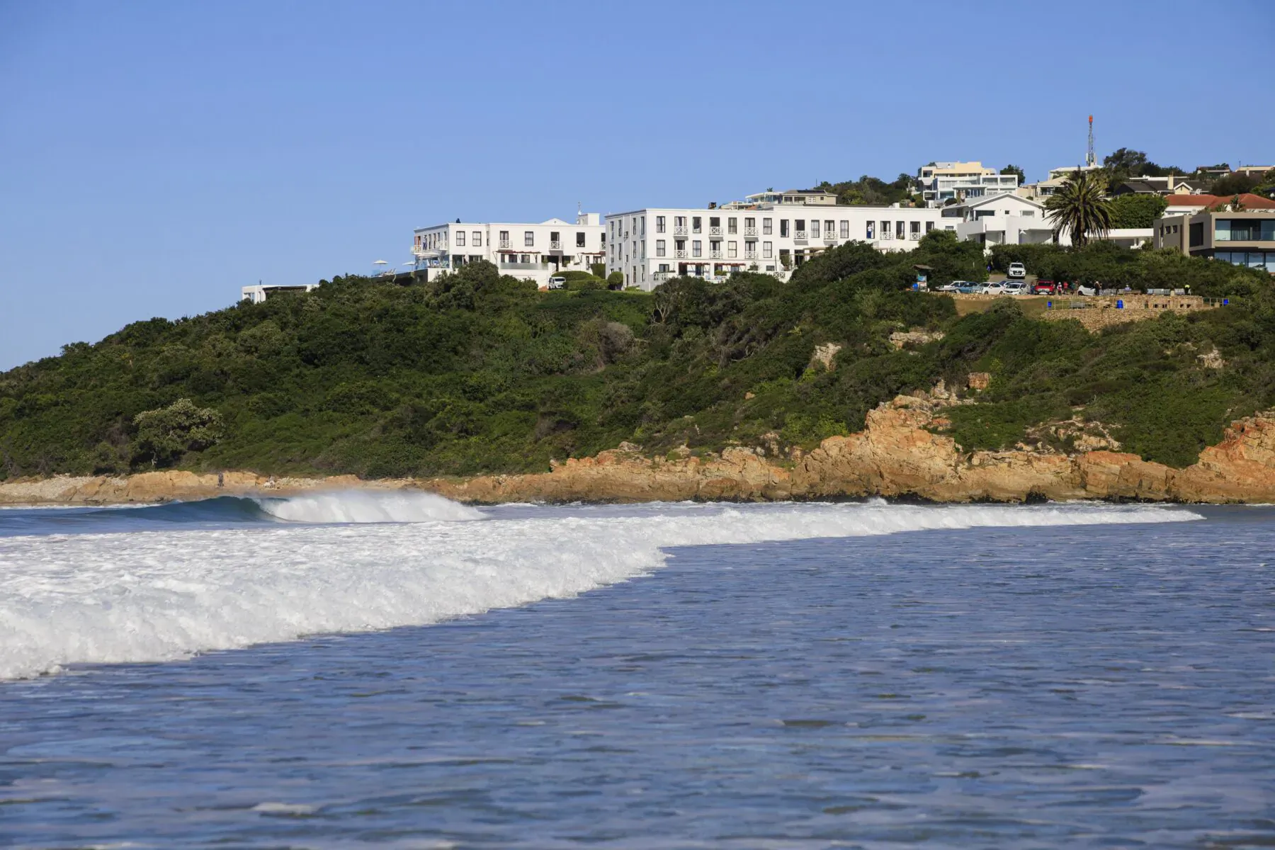 The Plettenberg Boutique Hotel on green hillside overlooking ocean waves, clear blue sky