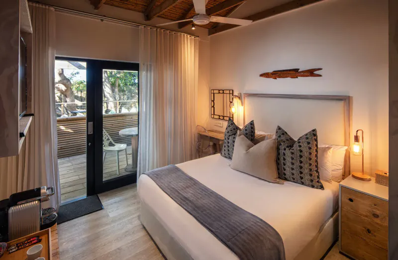 Seaside bungalow bedroom with king bed, pillows, lamps, open doors to deck with ocean view, ceiling fan, wood fish wall art.
