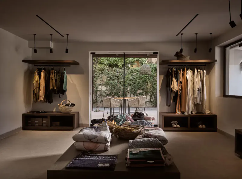 Luxury hotel boutique interior with wooden racks of hanging clothes, folded linens on table, and garden view through glass doors.