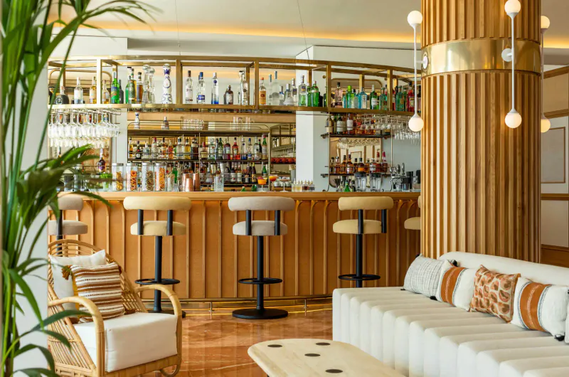 Elegant Hotel Riomar bar in Ibiza with wooden stools, gold shelves of liquor bottles, plants, and white sofas.