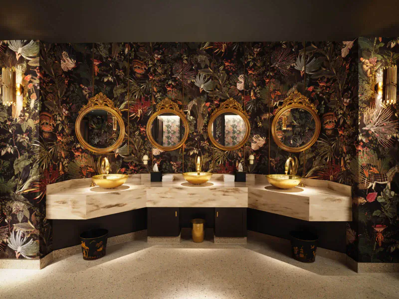 Luxurious washroom with three gold sinks, ornate round mirrors, and dark floral wallpaper in The Other House.