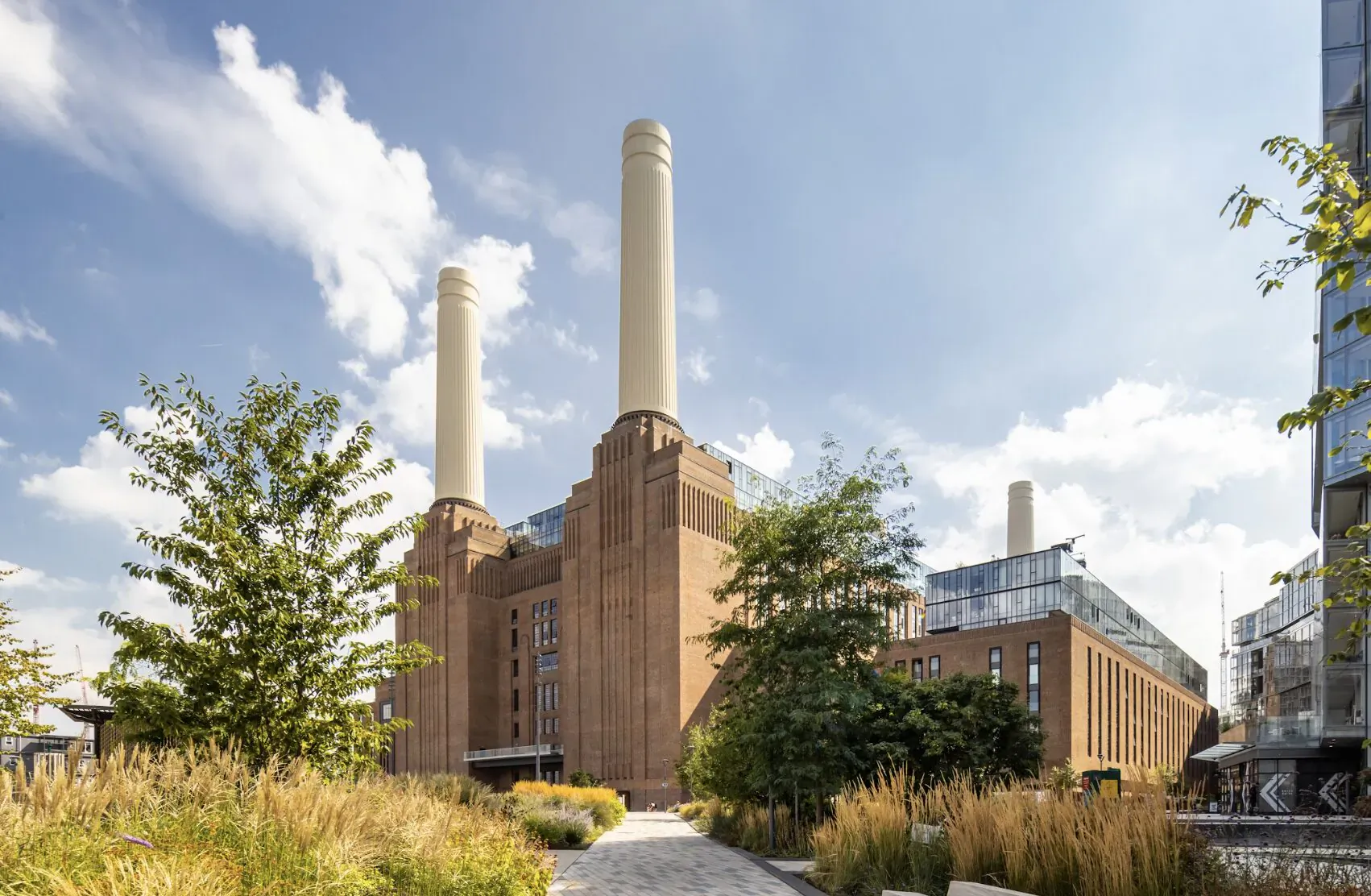 Battersea Power Station with iconic chimneys, surrounded by trees, grasses, and a riverside path on a sunny day.