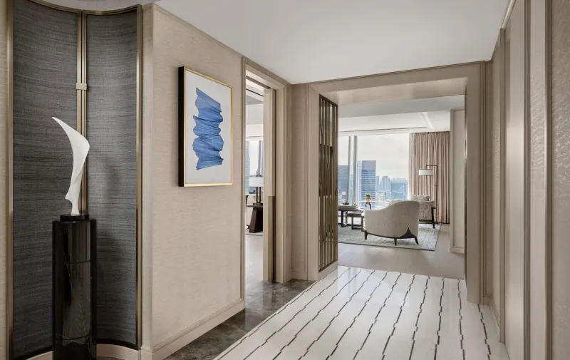 Luxurious Paiza Collection suite hallway with blue abstract painting, white sculpture, curved sofa, and city skyline view.