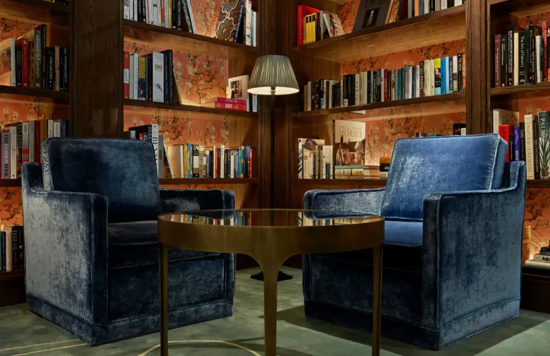 Cozy library lounge with blue velvet armchairs around a gold round table, surrounded by wooden bookshelves and a lamp.