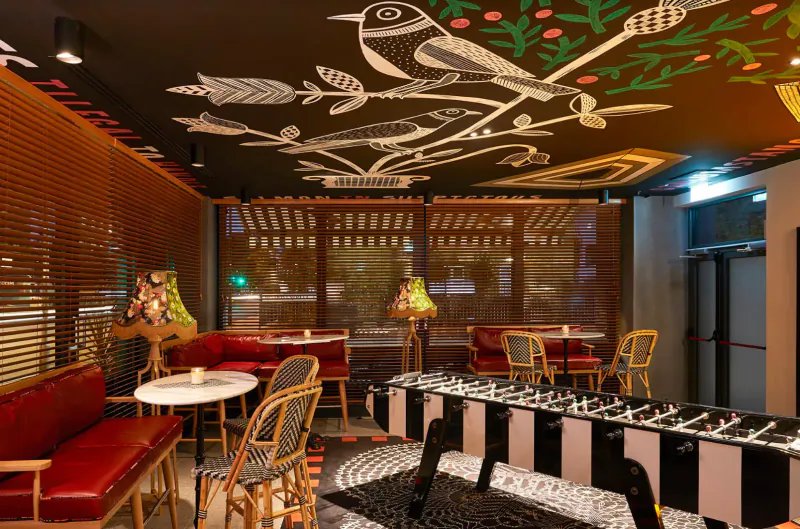 Cozy Mama Shelter bar interior with bird motifs on ceiling, red seating, foosball table, lamps, and large windows.