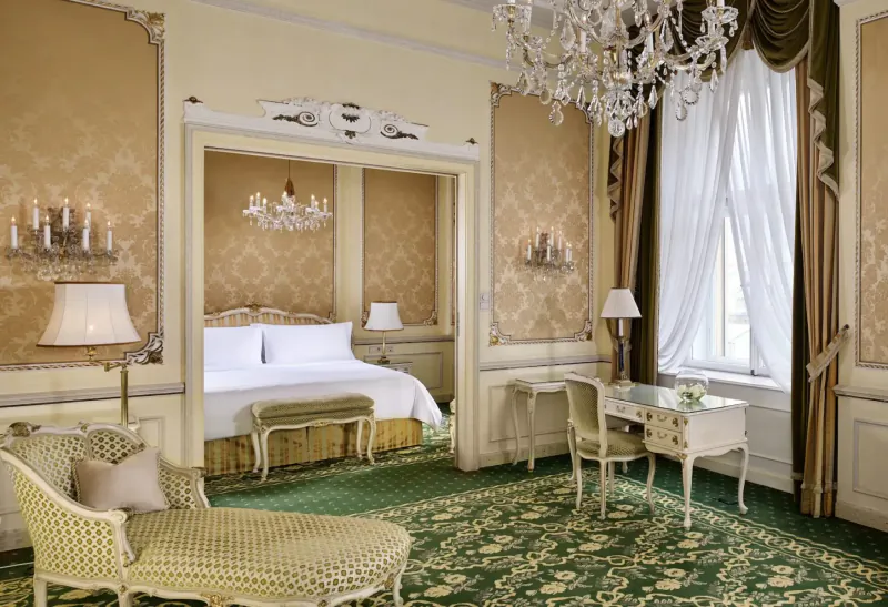 Luxurious Hotel Imperial Vienna suite with ornate gold walls, chandelier, king bed, desk, chaise, and green carpet.