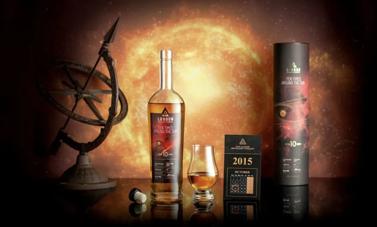 London Distillery Company 10-Year-Old single malt whisky bottle, glass with whisky, box marked 2015, beside armillary sphere on reflective surface.