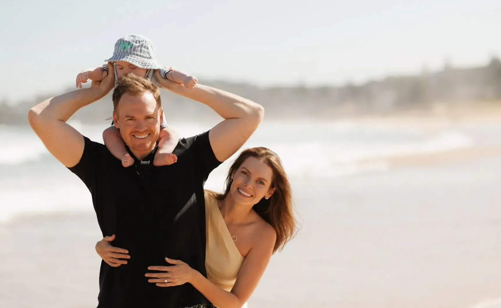 Jeremy holding baby atop his shoulders, embracing Elizabeth on sunny beach