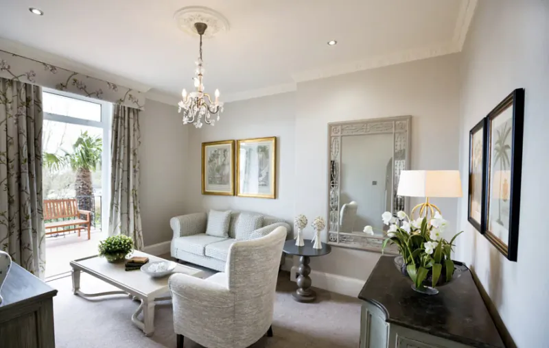 Elegant hotel sitting room with gray sofa and armchair, chandelier, large mirror, plants, and balcony view to palm trees and bay.