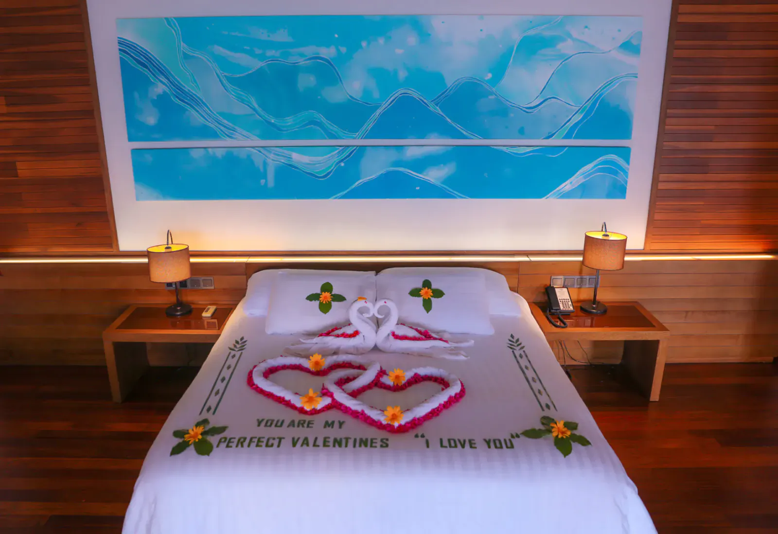 Romantic bed at Hideaway Beach Resort with swan towel hearts, frangipani, and 'You are my perfect Valentine I love you' on white sheets, blue artwork backdrop.