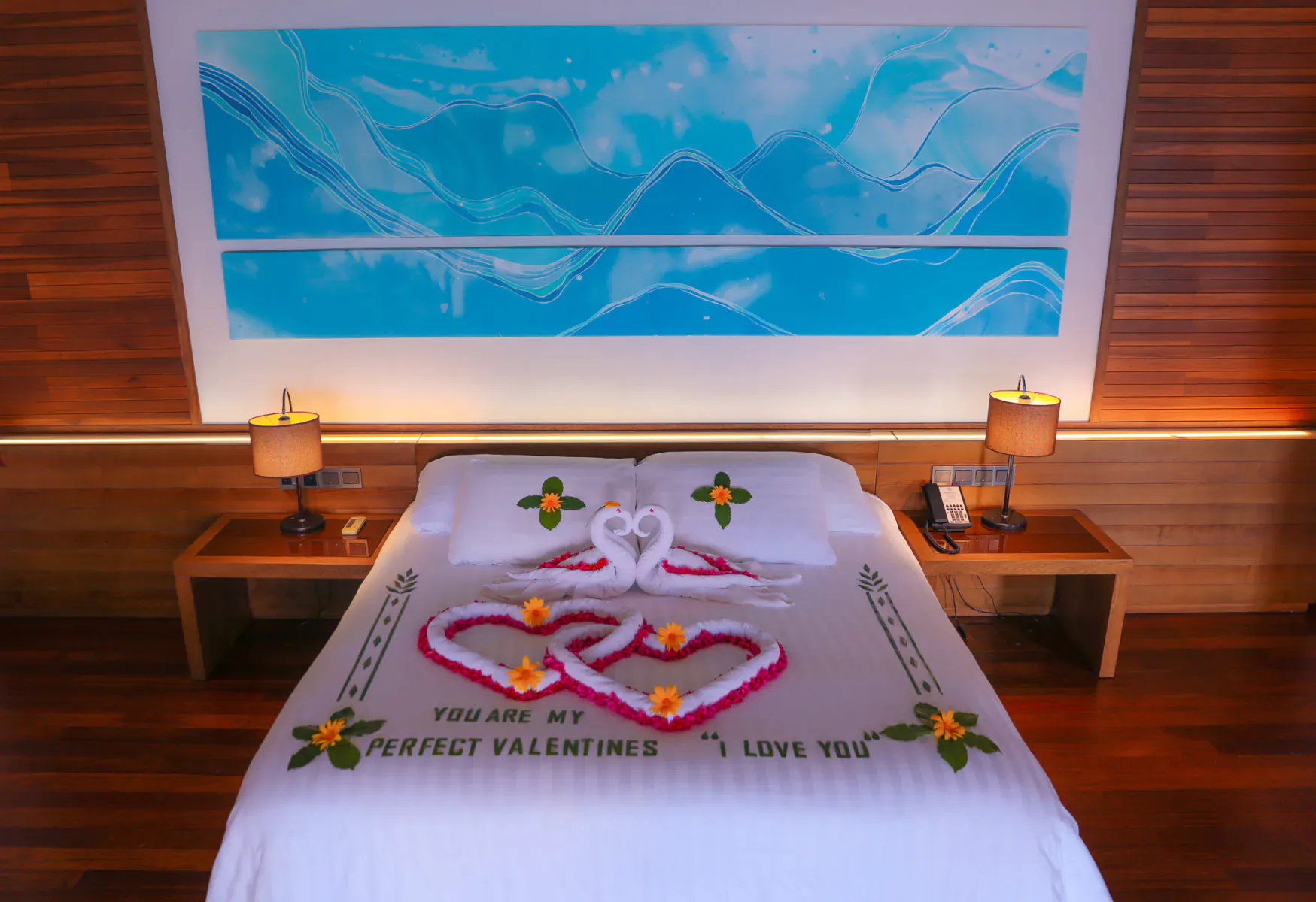 Romantic bed at Hideaway Beach Resort with swan towel hearts, frangipani, and 'You are my perfect Valentine I love you' on white sheets, blue artwork backdrop.