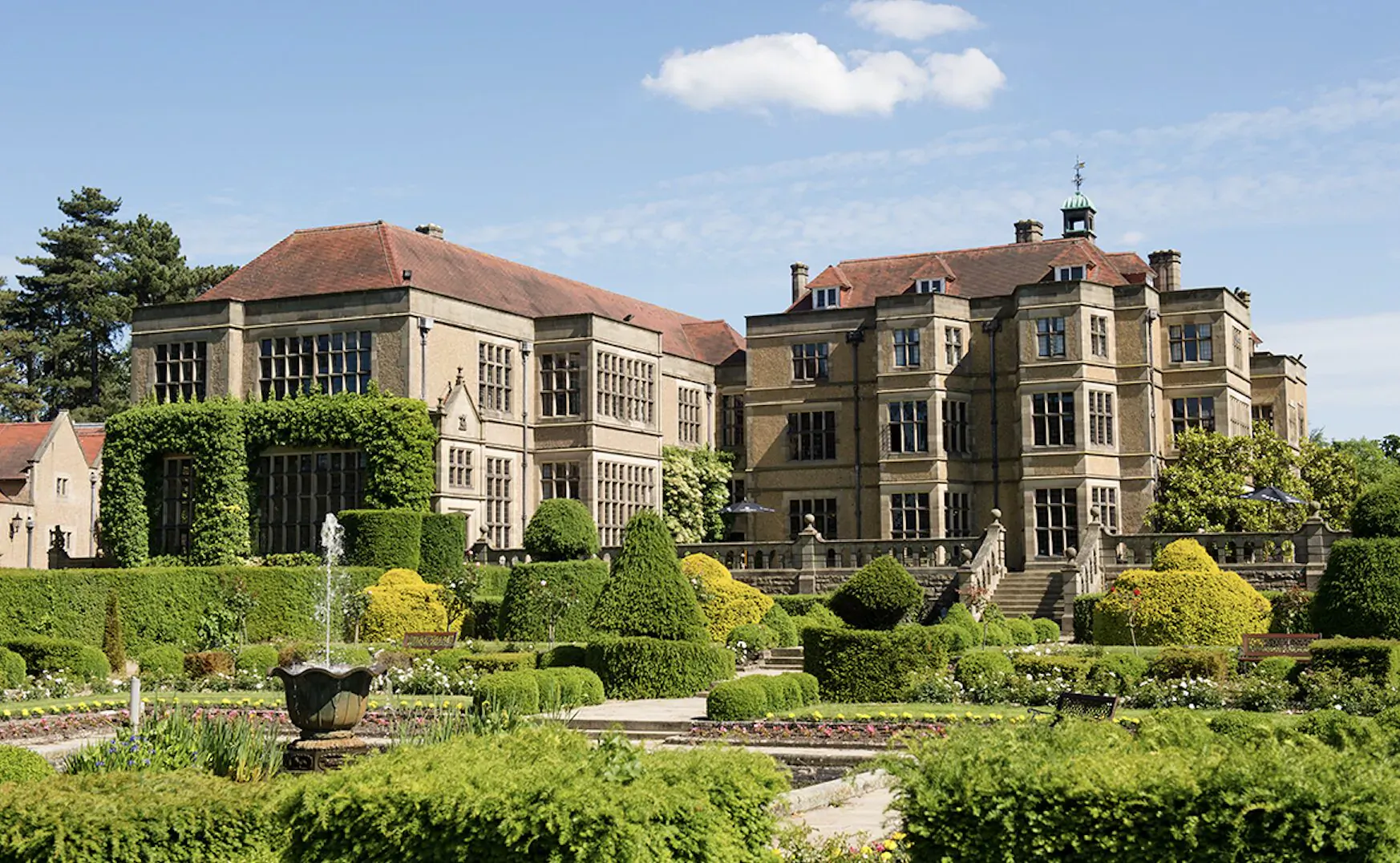Fanhams Hall Hotel, a grand Tudor-style mansion with manicured gardens, hedges, fountain, and trees under blue sky.