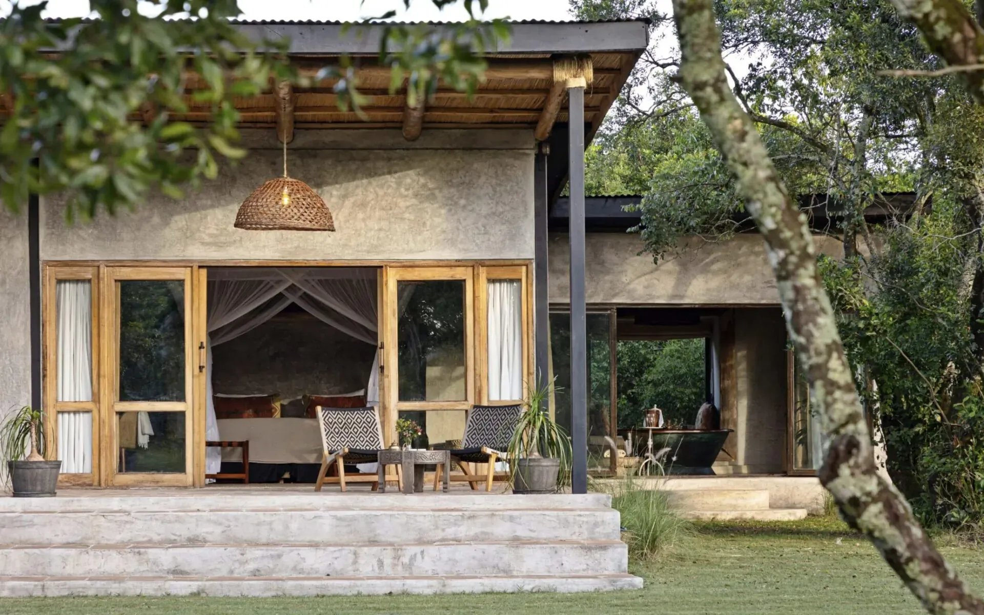 Eco-luxury house in Maasai Mara with open doors to bed, veranda chairs, plants, and lush trees.