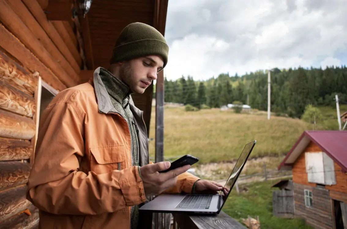 Man in green beanie and orange jacket works on laptop and phone on wooden cabin balcony overlooking forest meadow