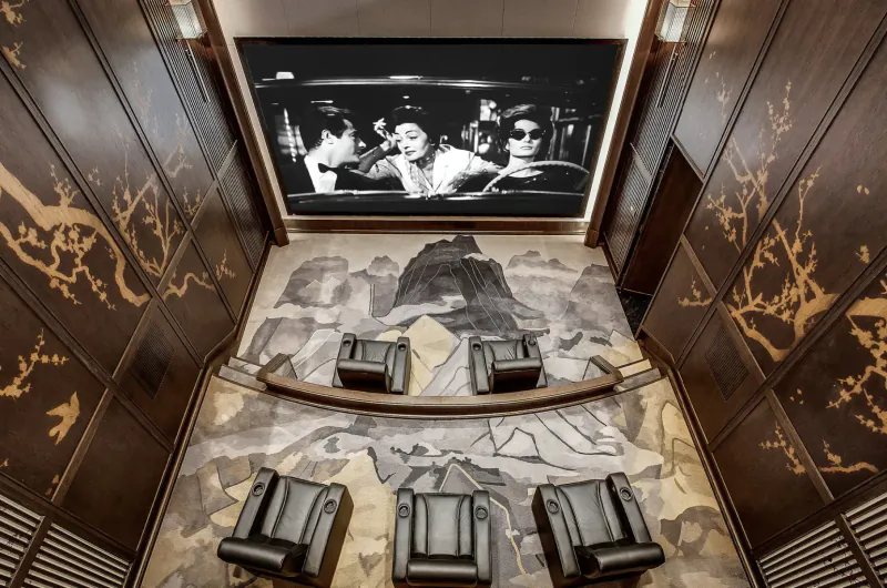 Top-down view of luxurious home theater with large black-and-white film on screen, leather chairs, ornate wood-paneled walls, and patterned floor.