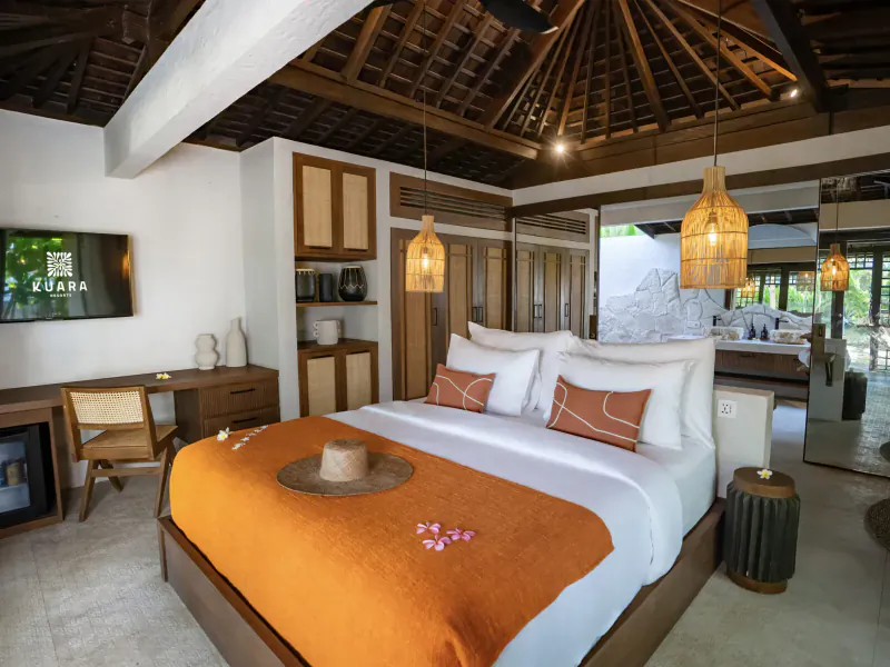 Luxurious bedroom in Kuara Lombok resort with king bed, orange throw, hat, lanterns, and garden view.