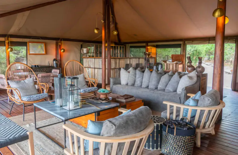 Luxurious safari tent lounge with gray sofa, wicker chairs, wooden tables, lanterns, and bush views.