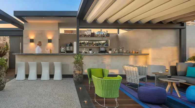 Rooftop bar at El Llorenç Parc de la Mar with bartender behind counter, modern green and white chairs, plants, sunset lighting.