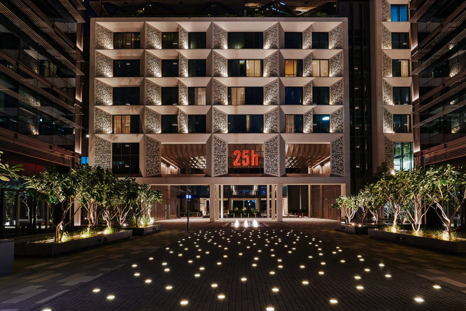25hours Hotel One Central in Dubai at night, modern facade with '25h' sign, palm trees, and illuminated courtyard paving