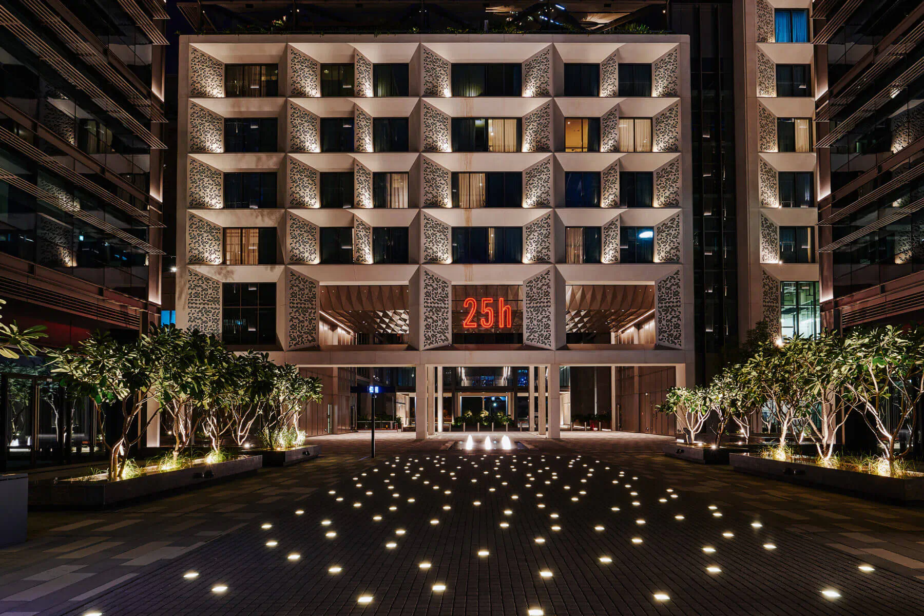25hours Hotel One Central in Dubai at night, modern facade with '25h' sign, palm trees, and illuminated courtyard paving