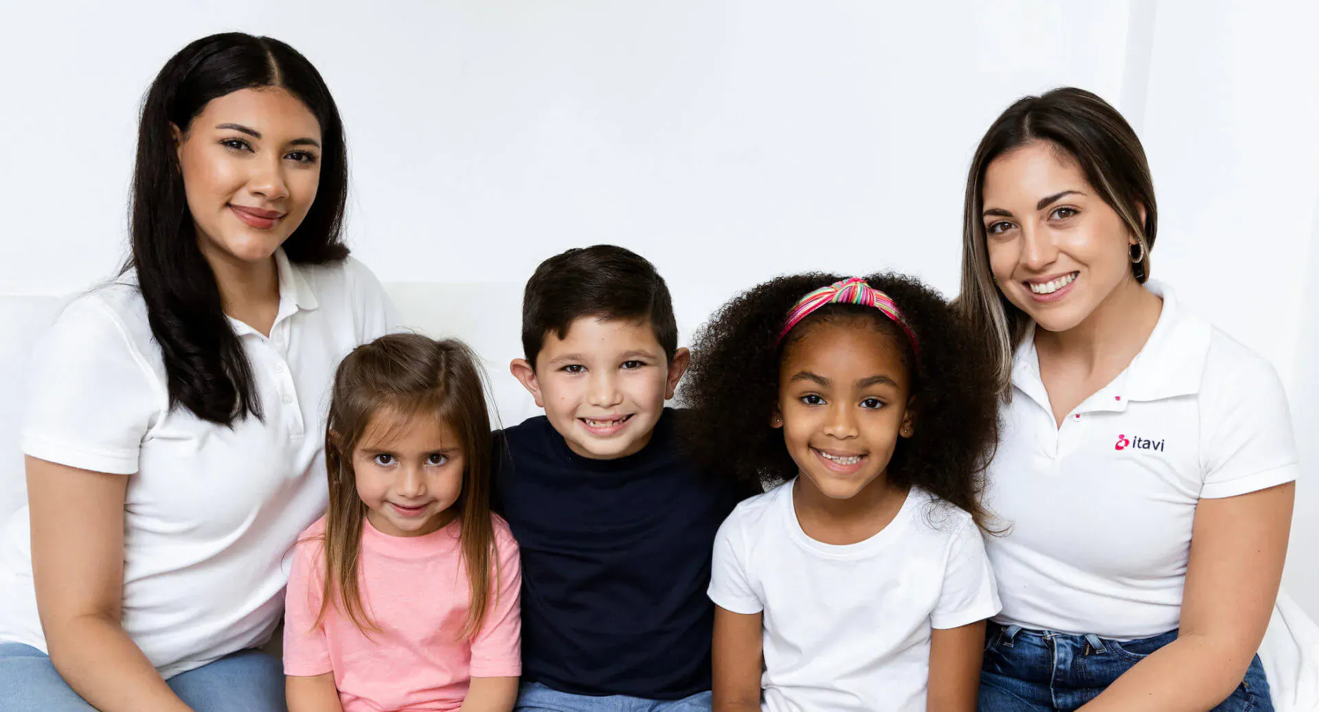 Two smiling women in white polo shirts with three young children posing happily on white background