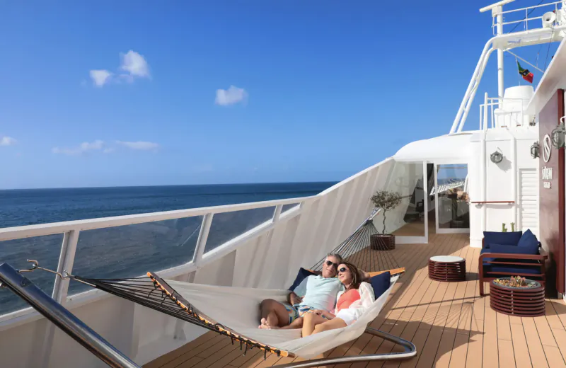 Couple relaxing in white hammock on luxury yacht deck, overlooking blue ocean under clear sky.
