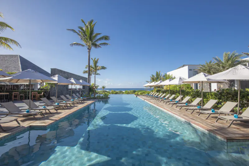 Infinity pool at Anantara Iko Mauritius Resort with loungers, white umbrellas, palm trees, and ocean view under blue sky.